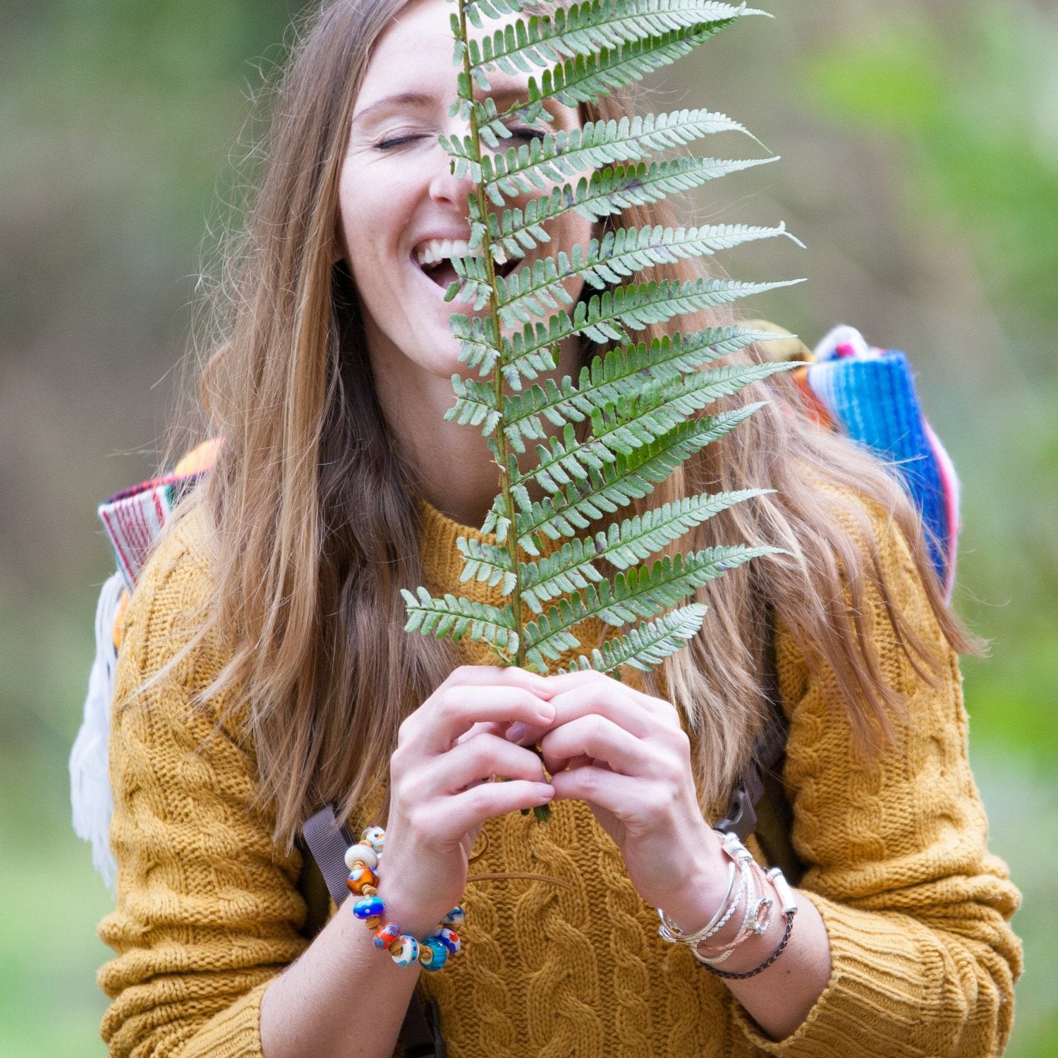 Woman in a rainforest holding a fern, wearing a yellow knitted jumper and a back pack with beaded jewellery.