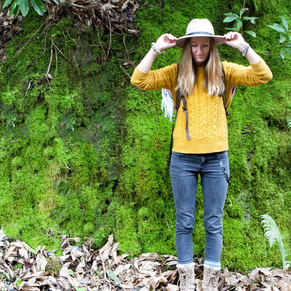 Woman standing in the green mossy woods wearing jeans and yellow knitted jumper with a white hat.