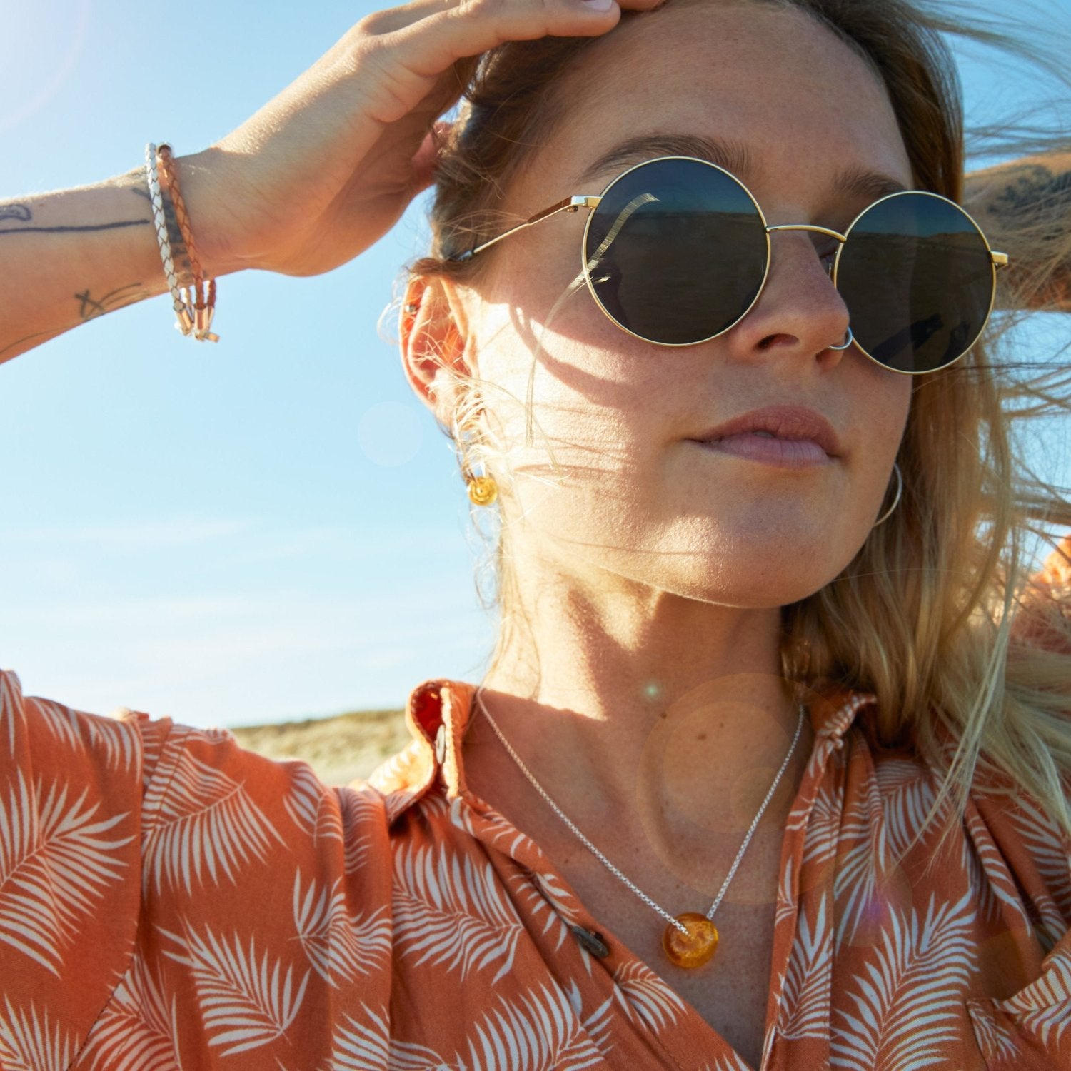 Girl wearing sunglasses and silver jewellery with a orange printed shirt on at the beach.