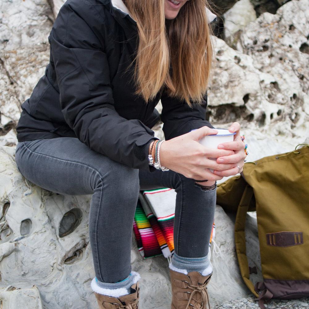 Woman sitting at the beach in winter with back pack holding a cup of tea,