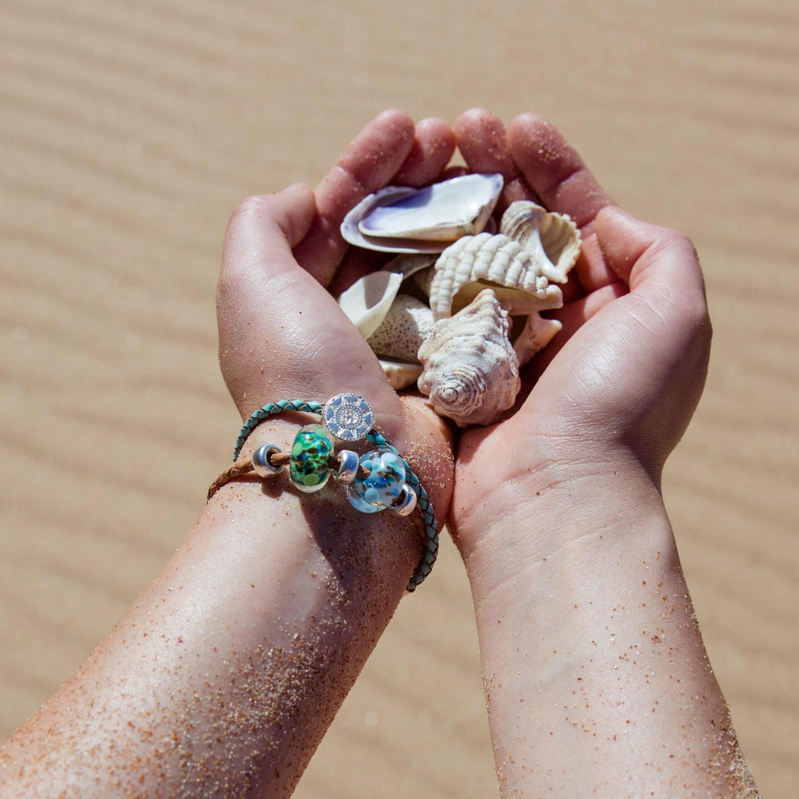 Hands holding shells on the beach while wearing beaded bracelets.