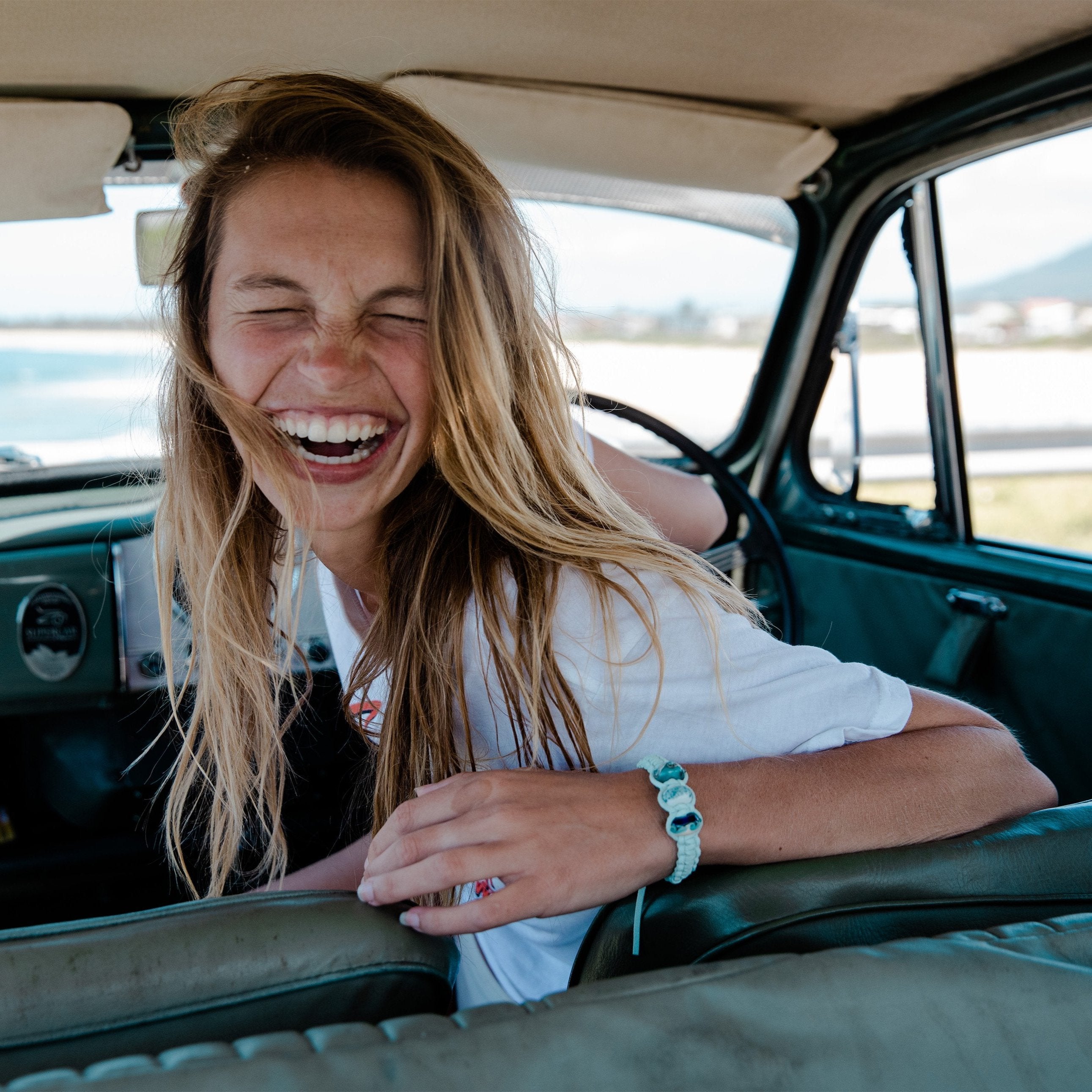 Woman sitting in her car, wearing a white t-shirt and braided bracelet.