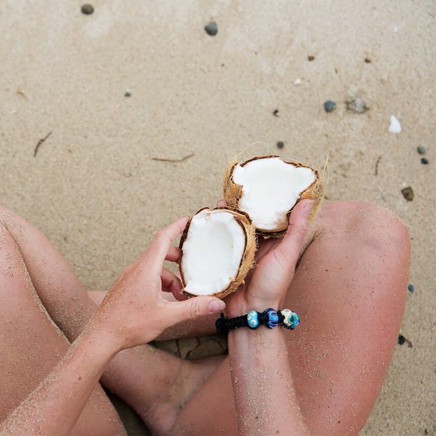 Person sitting on the beach cross legged, holding a coconut wearing a black cord bead bracelet.