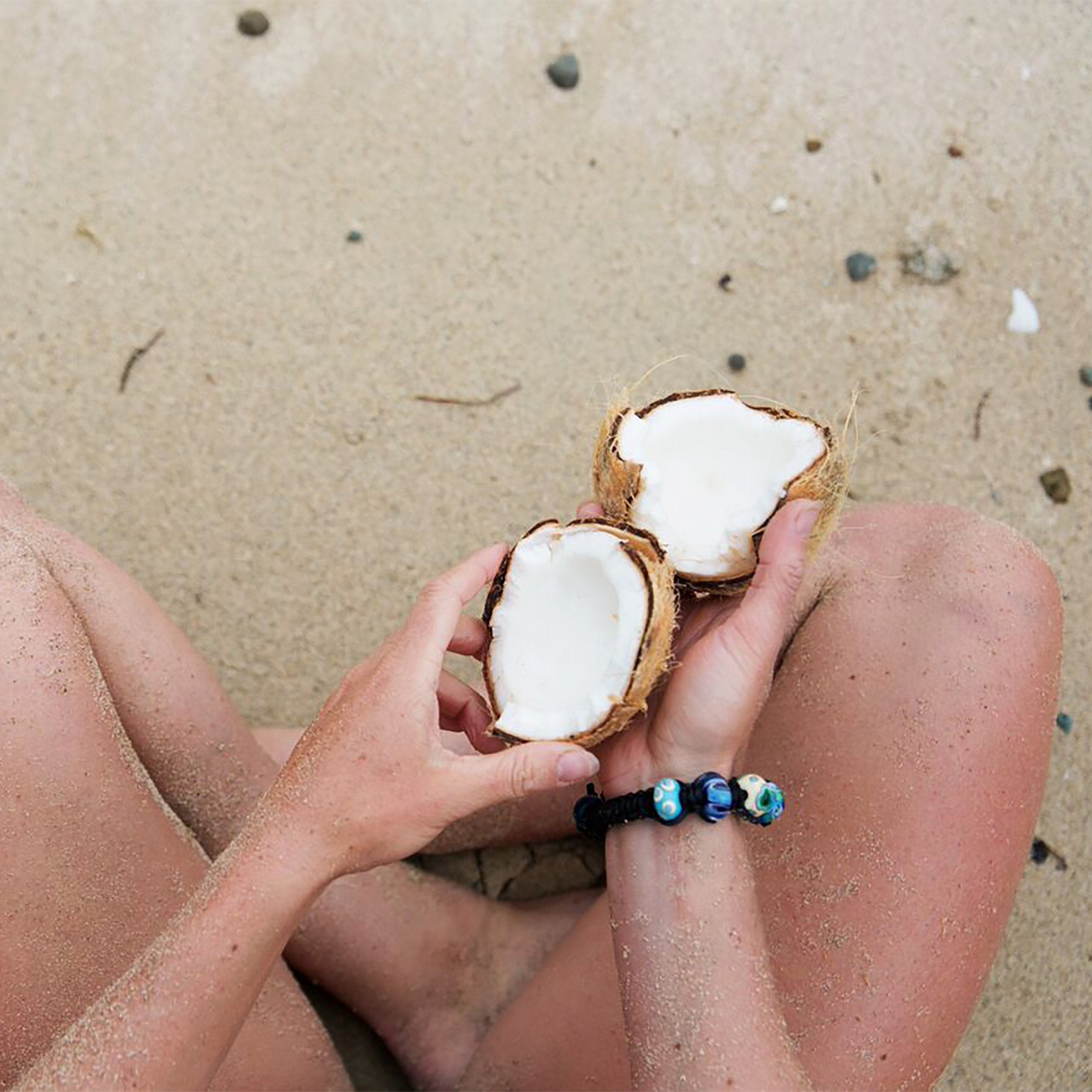 Person sitting on the beach cross legged, holding a coconut wearing a black cord bead bracelet.