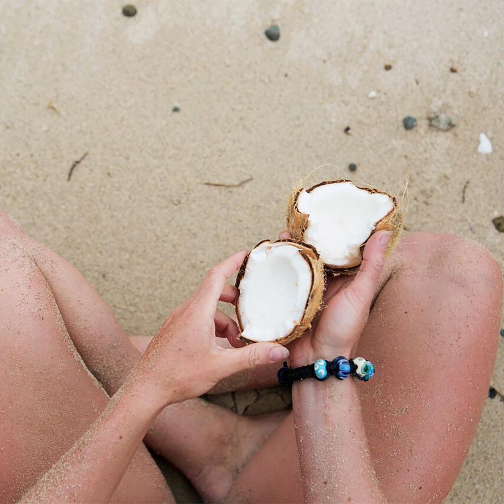 Woman wearing black cord bead bracelet and a coconut on the sand.