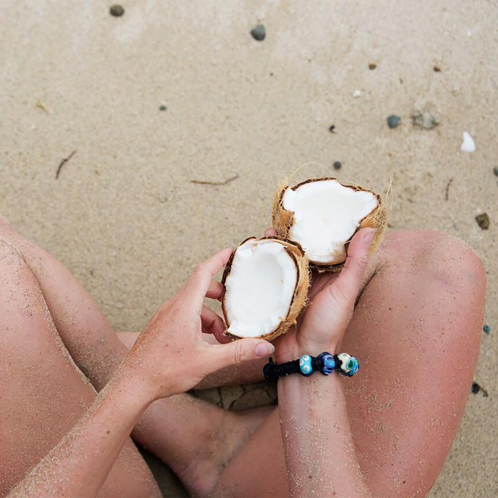 Person sitting crossed legged on beach, holding a coconut wearing a black cord beaded bracelet.