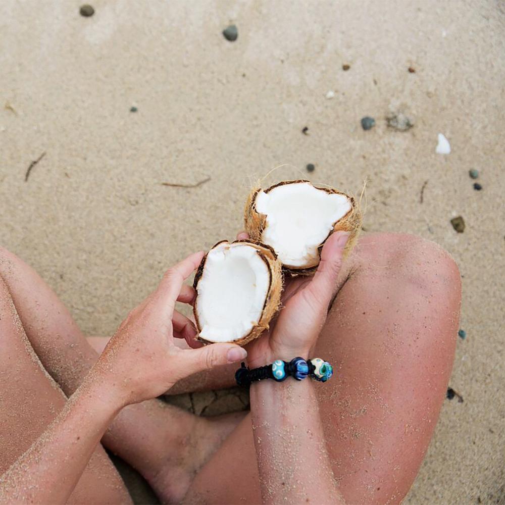 Person sitting on the beach holding a cracked coconut, wearing a black cord bead bracelet.