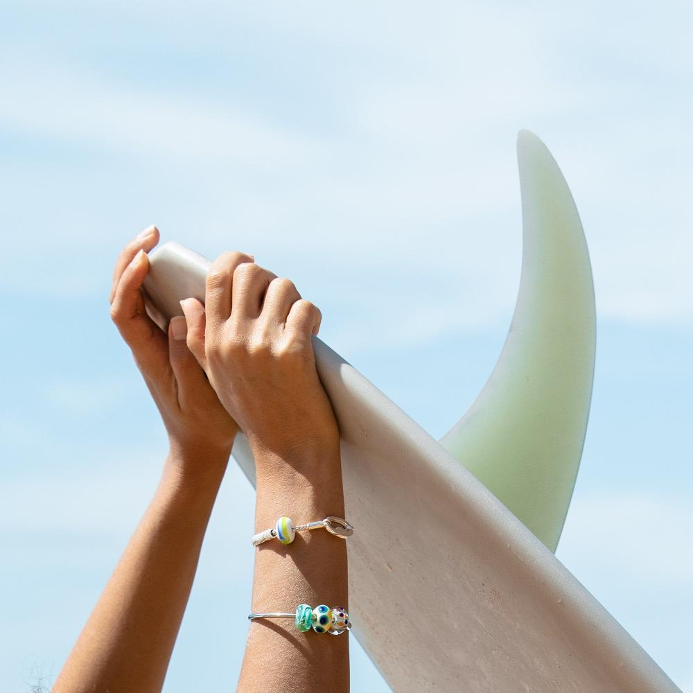 Surfer wearing silver beaded bangle and bracelet while holding surfboard with single fin.