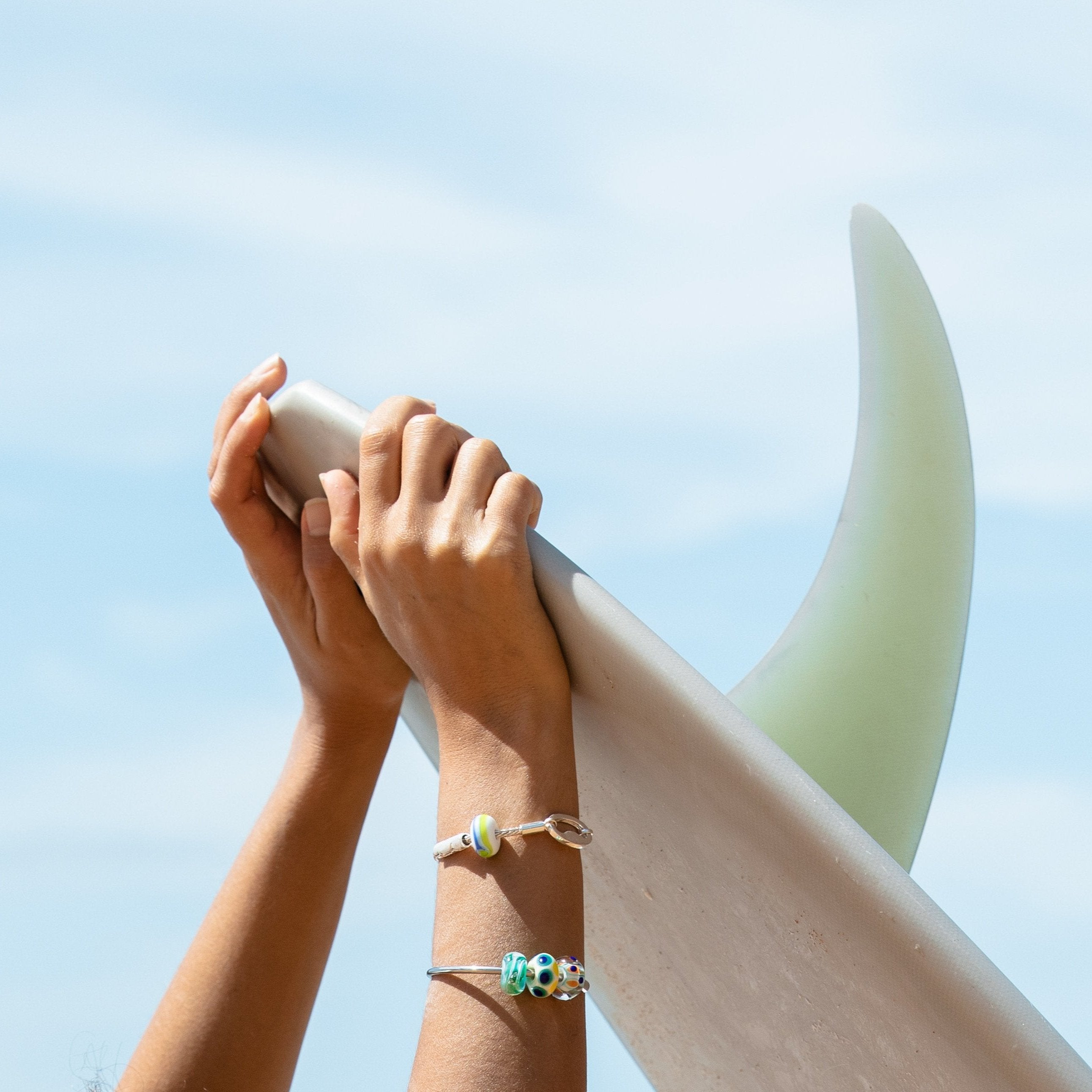 Hands holding up surfboard wearing Nalu silver bracelets and bangle with glass travel charms.