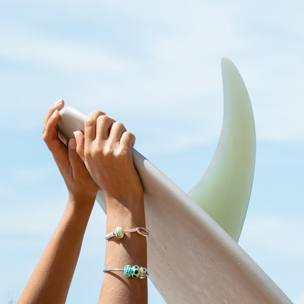 Hands holding surfboard with fin wearing silver bracelet and bangle.