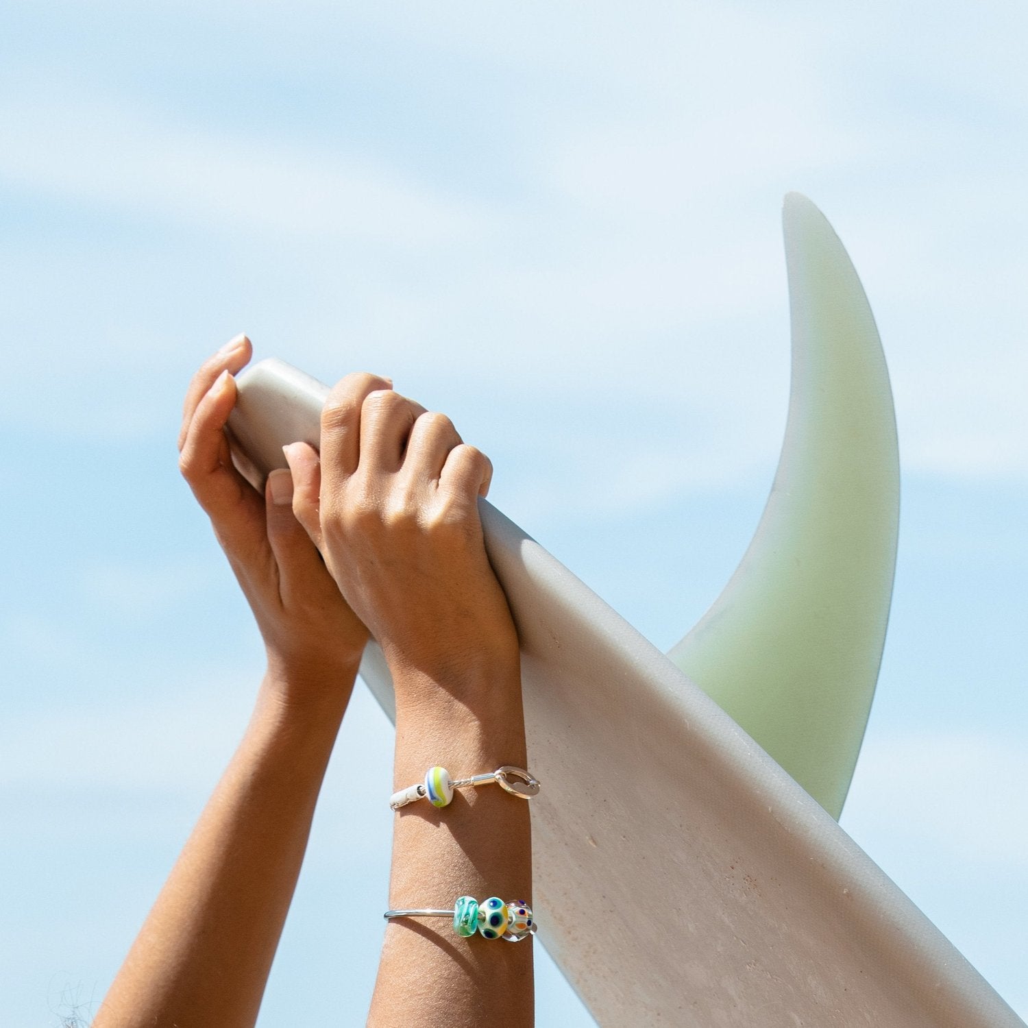 Surfer wearing silver beaded bangle and bracelet while holding surfboard with single fin.