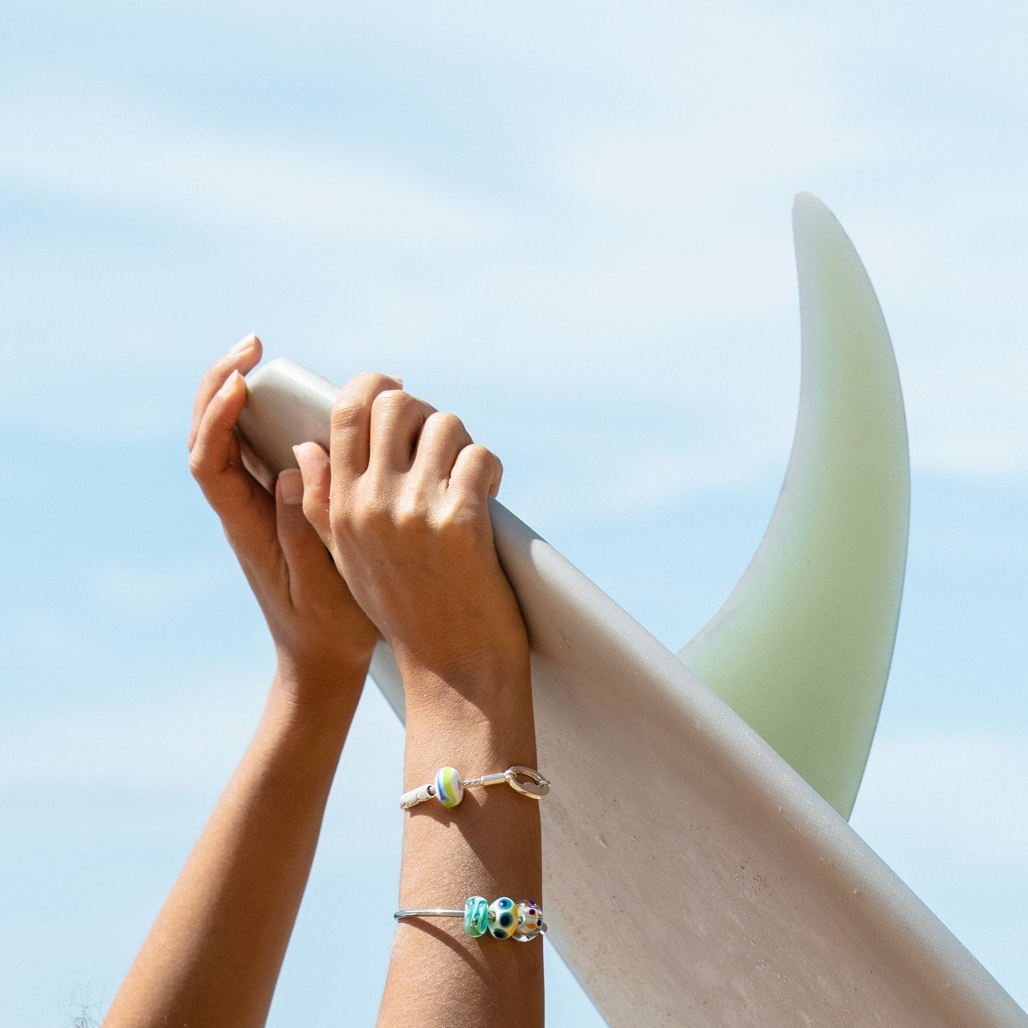 Surfer wearing silver beaded bangle and bracelet while holding surfboard with single fin.