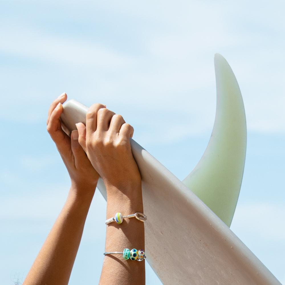 Surfer wearing silver beaded bangle and bracelet while holding surfboard with single fin.