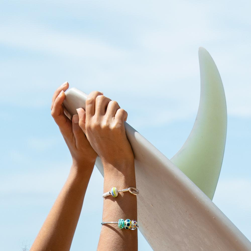 Surfer wearing silver beaded bangle and bracelet while holding surfboard with single fin.