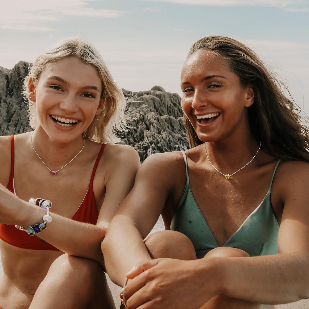 Two ladies sitting on the beach at Croyde Bay wearing bikinis and beach sand pebble necklaces jewellery.