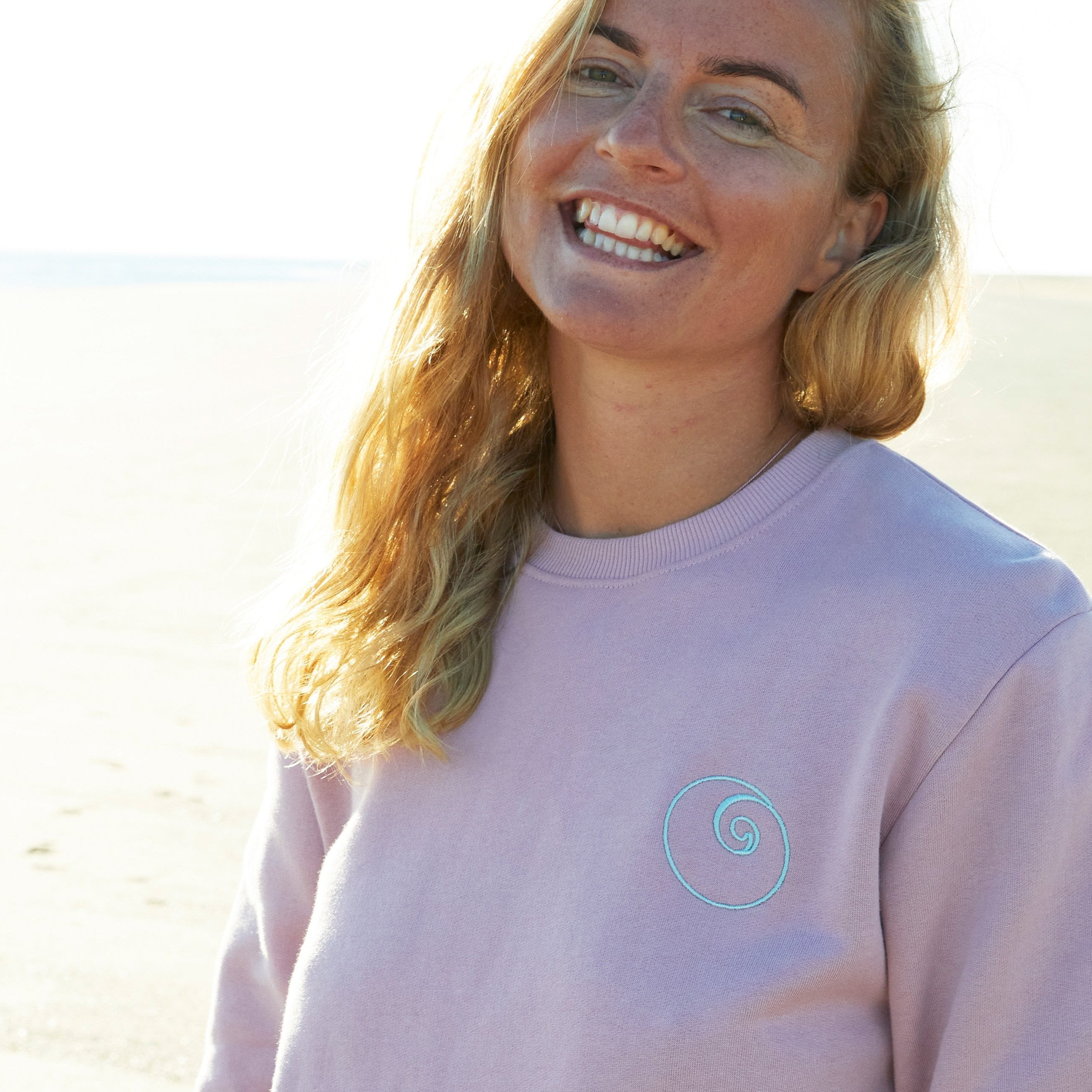 Woman with long blonde hair wearing a pink sweatshirt at the beach