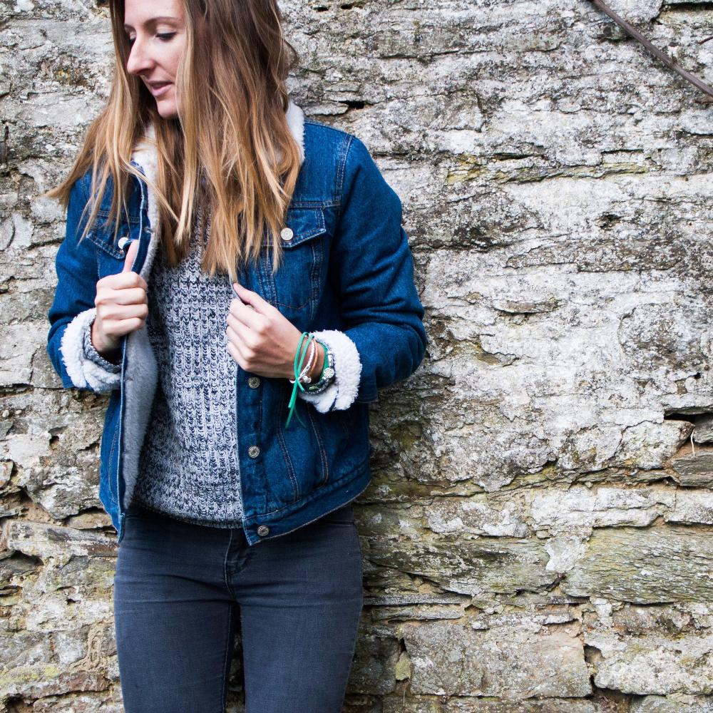 Woman standing against brick wall wearing denim jacket and jeans with bracelet stacks.