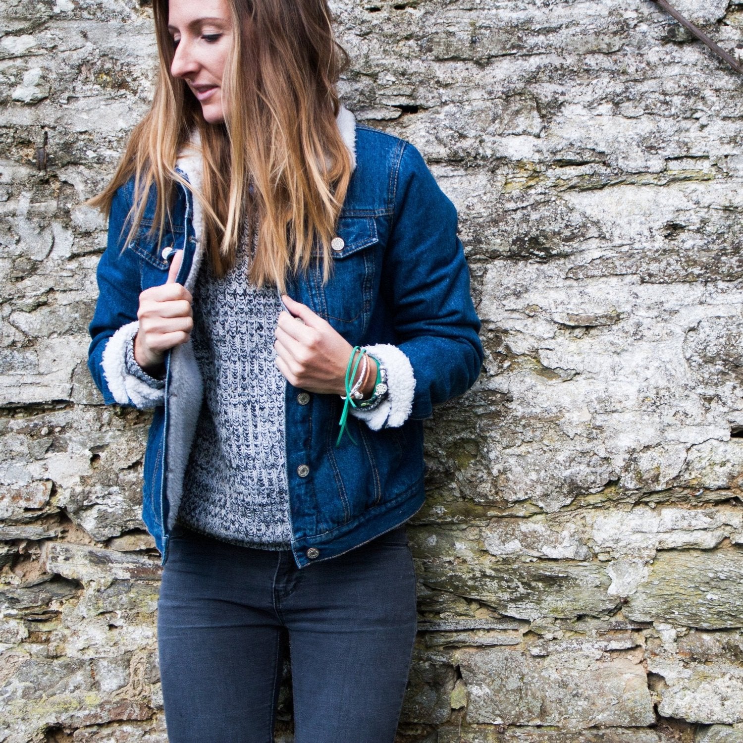 Woman standing against the wall wearing a denim jacket and bracelets.