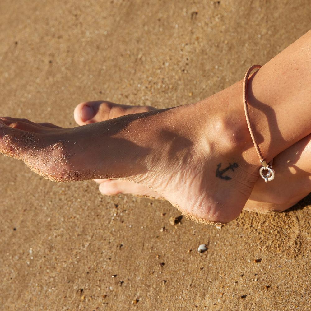 Person on the beach with anchor tattoo on ankle, feet crossed with leather and silver anklet.