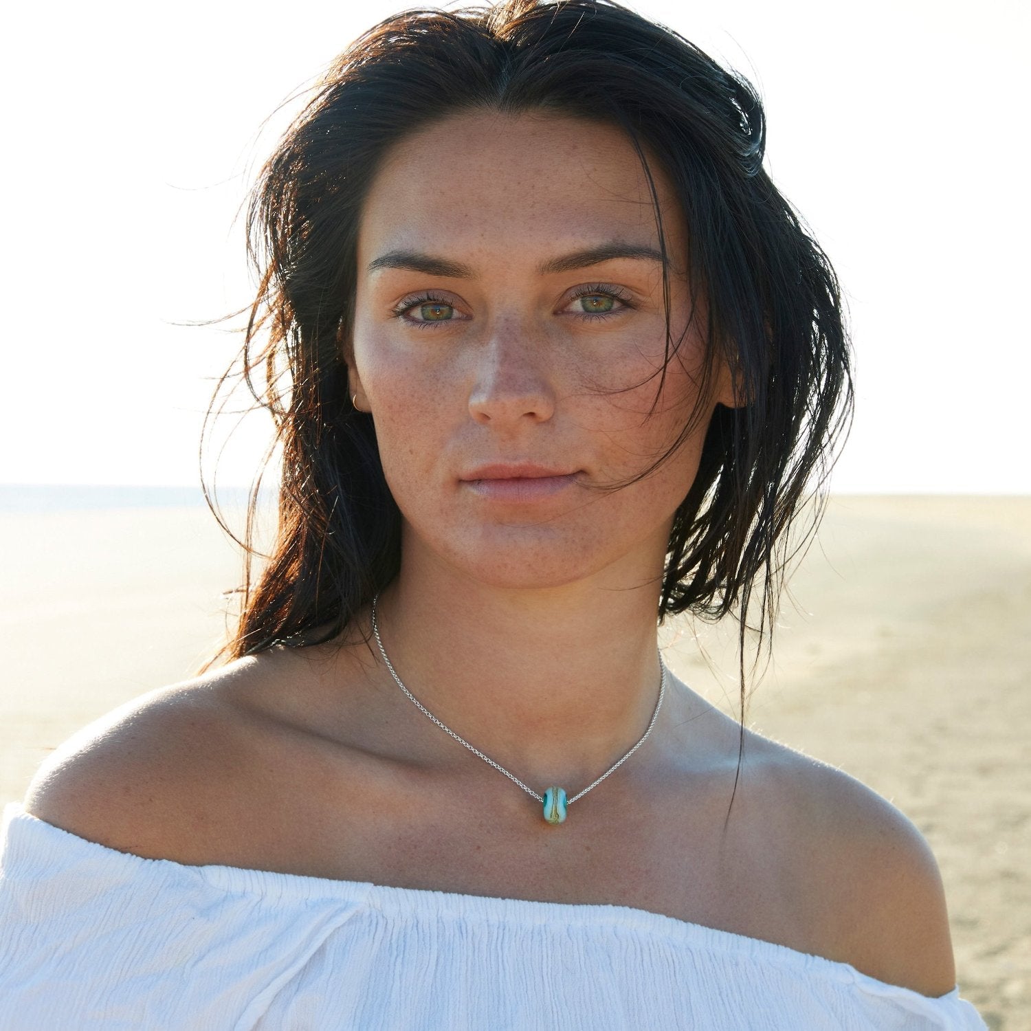 Dark haired woman standing on the beach wearing a silver chain bed necklace.