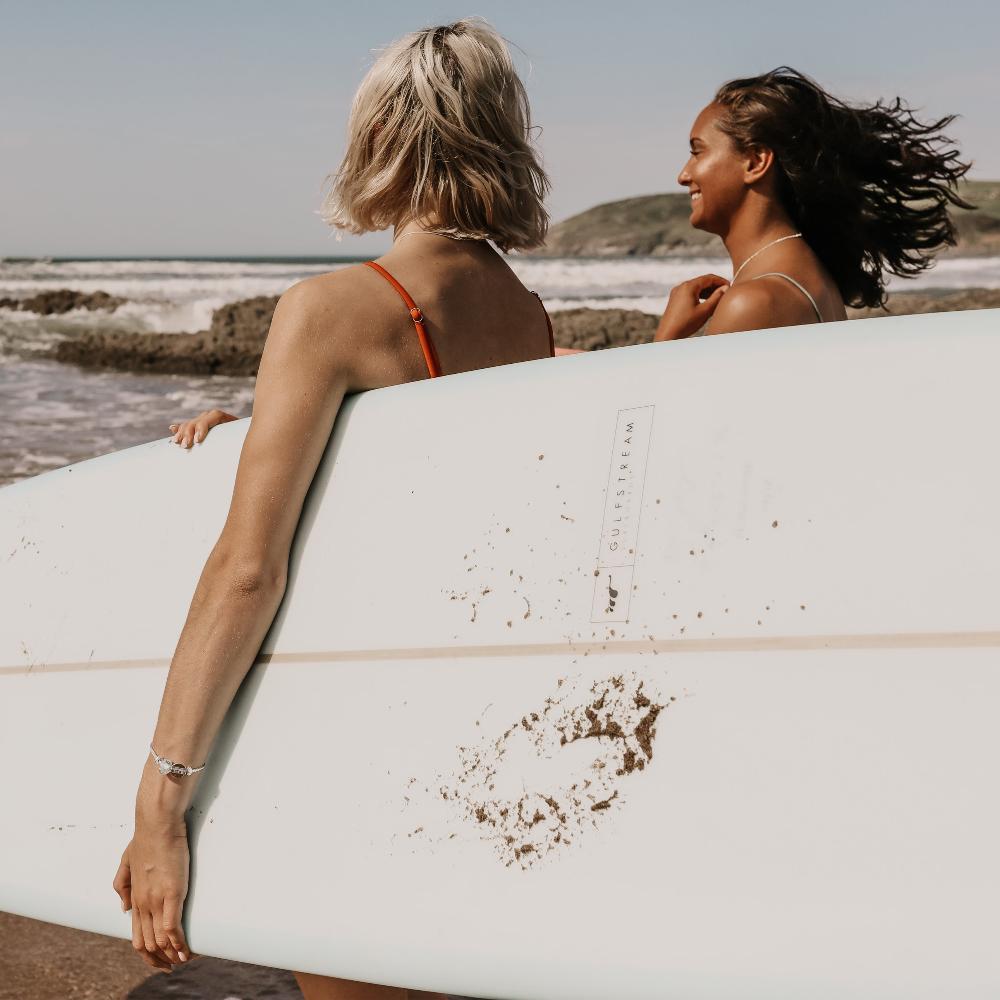 Girls carrying surfboard on beach walking into sea.