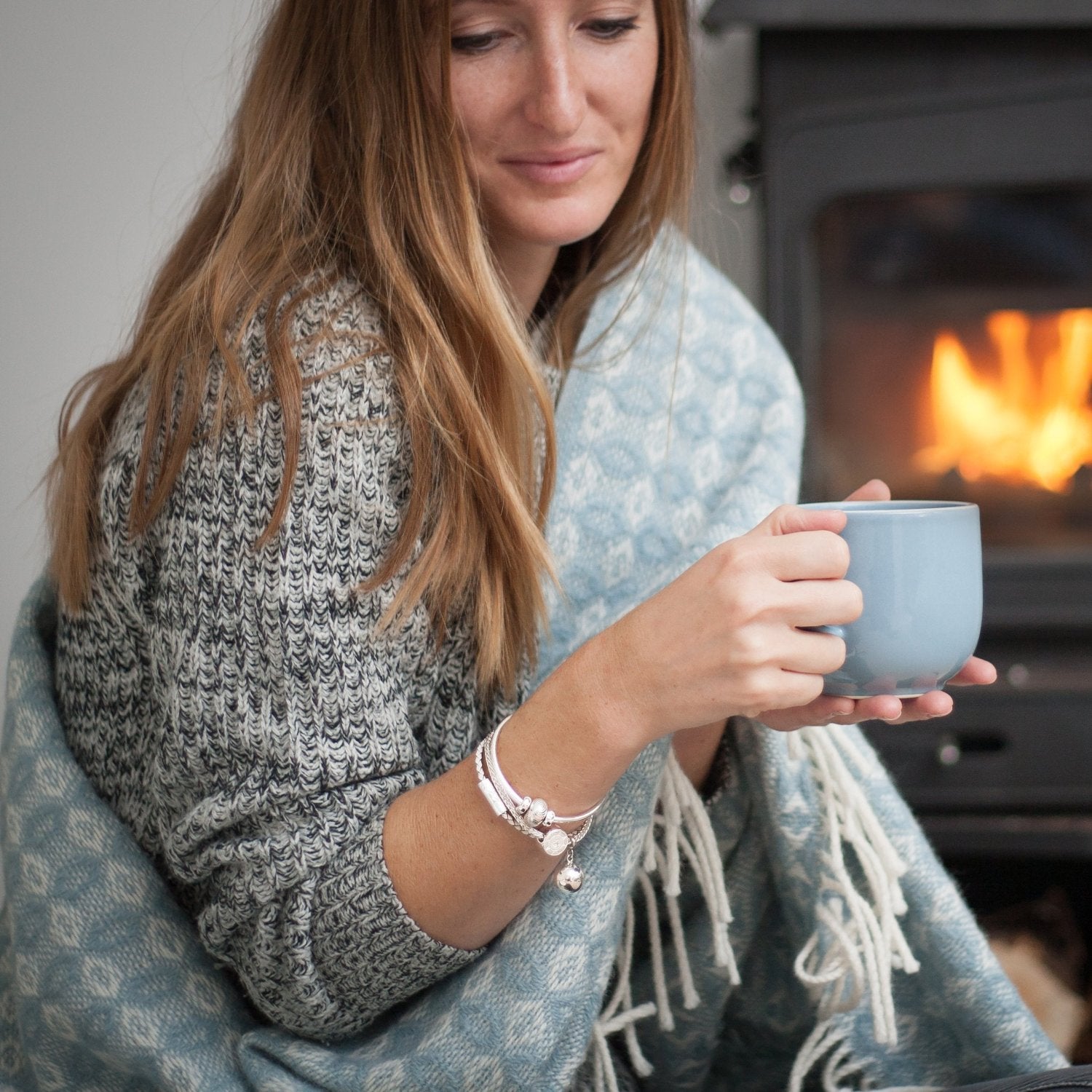 Woman sitting in front of a log burner, holding a cup of tea with a blanket over her shoulder. Wearing layers of silver bracelets.