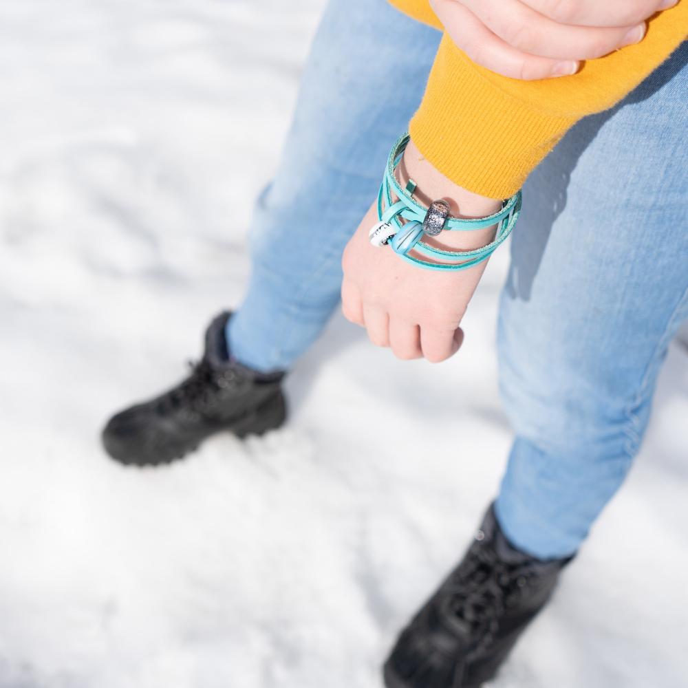 Person standing on the snow, wearing jeans and black boots with a turquoise wrap beaded bracelet.