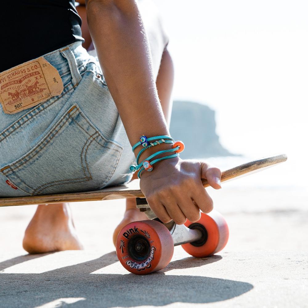 Girl in denim shorts sitting on a skateboard wearing a turquoise leather wrap bracelet with orange beads on it.