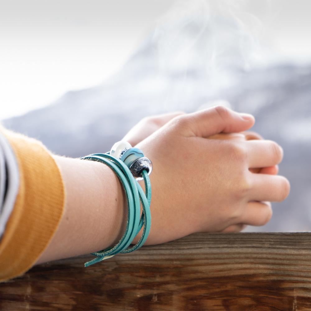 Hands holding a cup of steaming coffee, wearing a turquoise leather, glass bead bracelet. 