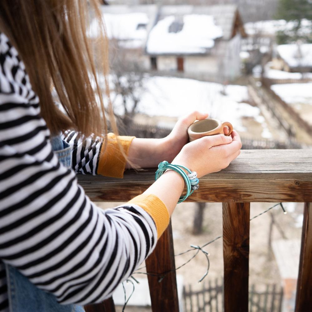 Hands holding a cup of steaming coffee, wearing a turquoise leather, glass bead bracelet.