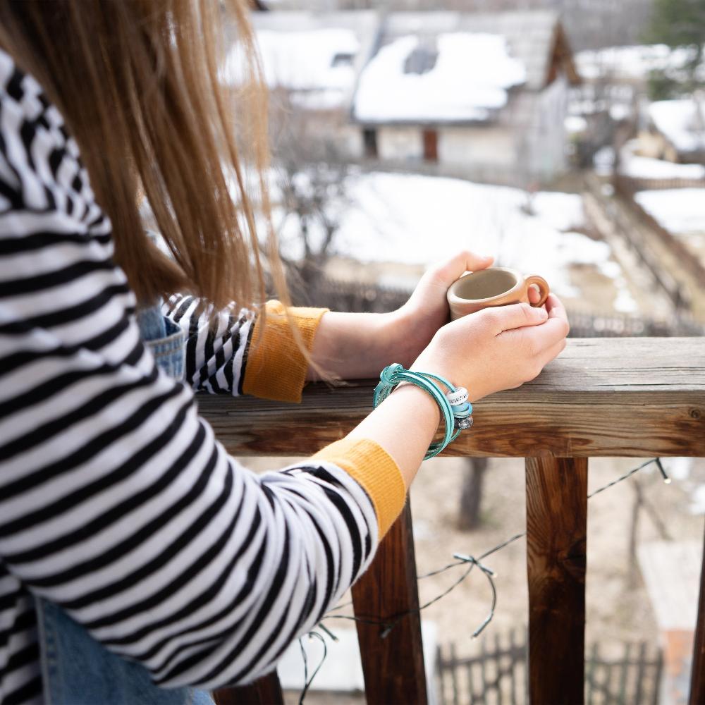 Hands holding a cup of steaming coffee, wearing a turquoise leather, glass bead bracelet. 