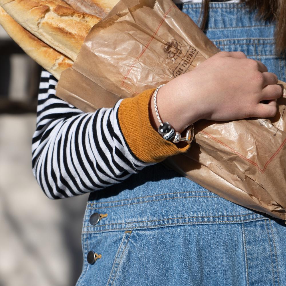 A girl carrying loaves of bread in denim dungarees wearing a white plaited bracelet with black and white glass beads.